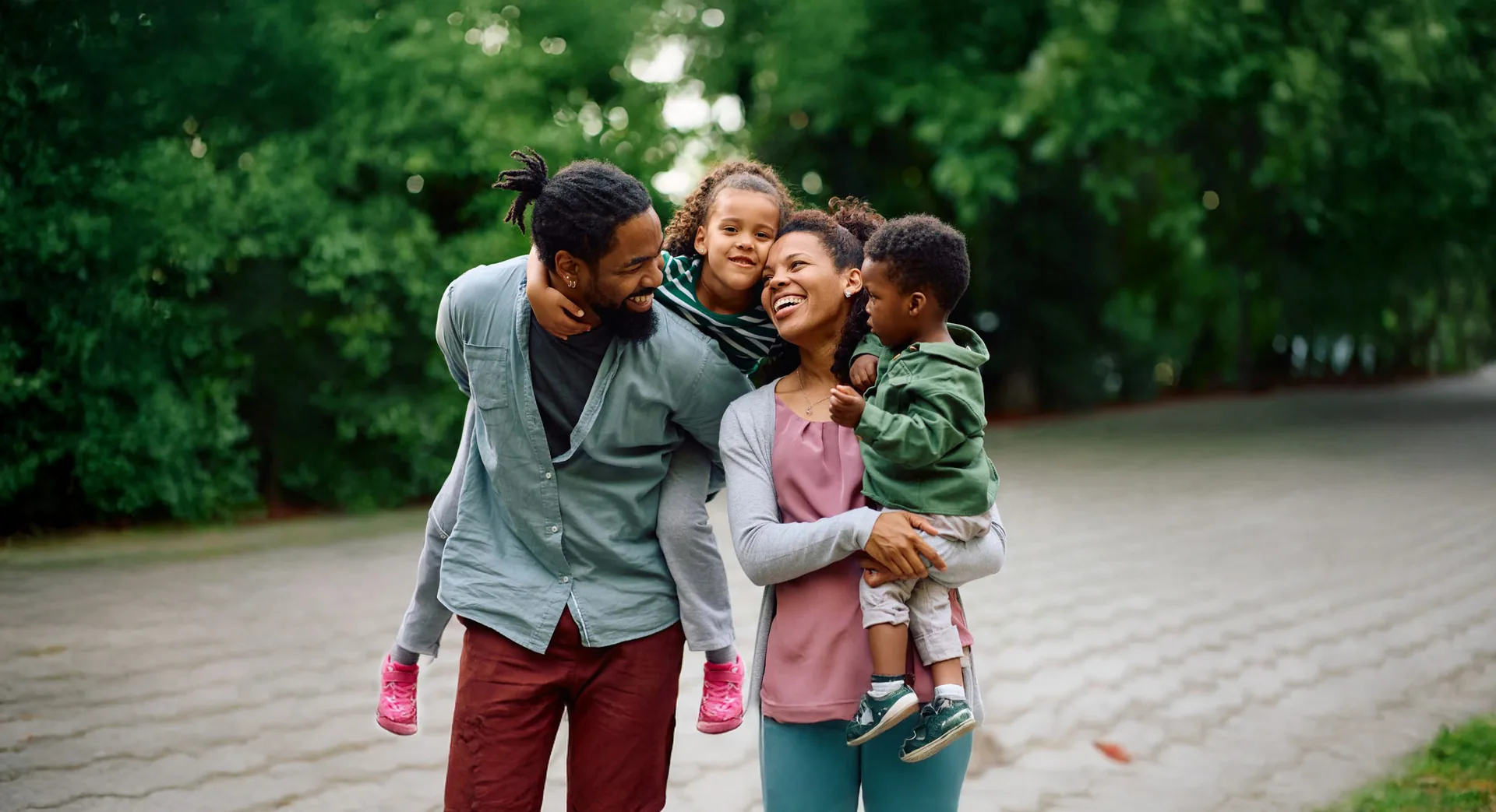 A family of four walking together along a tree-lined path, the parents each carrying a young child and all of them laughing