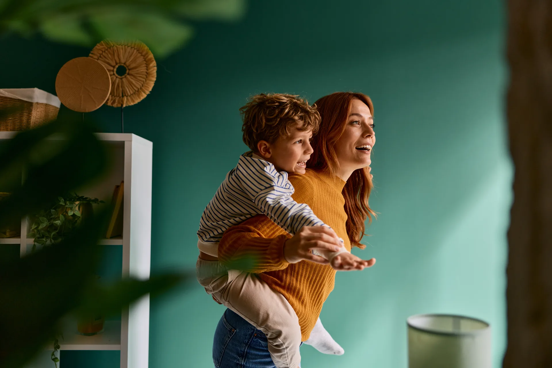A mother giving her young son a piggyback ride in a bright living room, both of them laughing